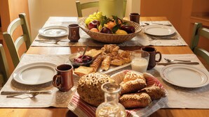 Una mesa de desayuno servida con platos, cubiertos, un bol de fruta, pan, pasteles y una botella de mantequilla.