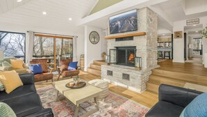 A spacious living room with a stone fireplace, comfortable seating, and large windows offering a view of the outdoors. - Mountain Views, Hot Tub, Impeccable Design (Wintergreen Resort)