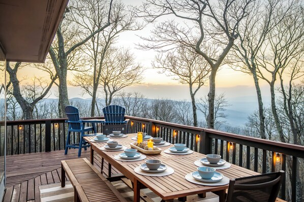 A wooden deck with a table set for dining, surrounded by chairs, overlooking a forested landscape. - Mountain Views, Hot Tub, Impeccable Design (Wintergreen Resort)