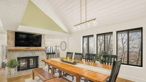 A dining area with a wooden table, chairs, a bench, a fireplace, a TV mounted on a stone wall, and large windows with a view of trees. - Mountain Views, Hot Tub, Impeccable Design (Wintergreen Resort)