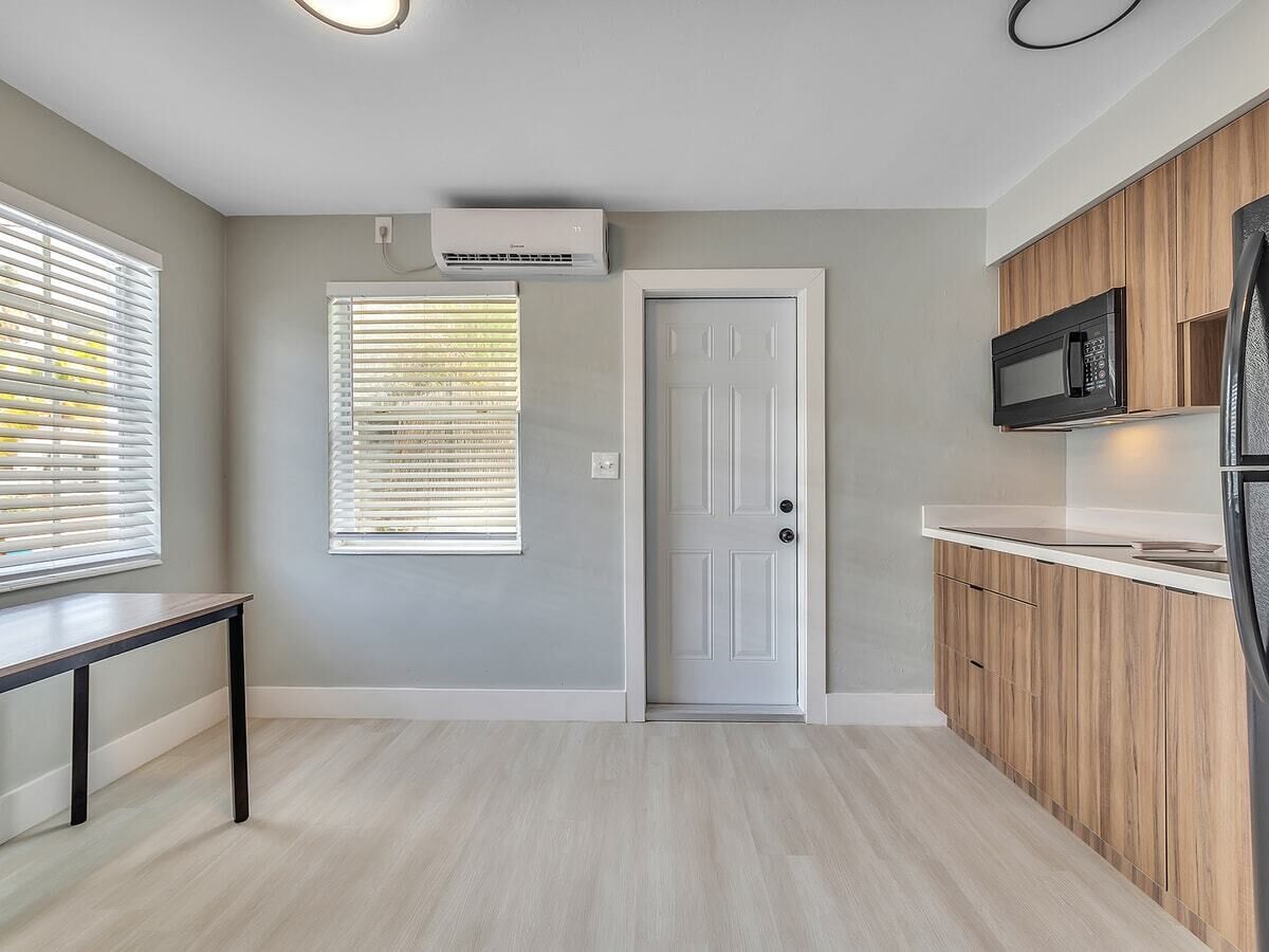 A modern kitchen with a white door, wooden cabinets, a black microwave, and an air conditioning unit.