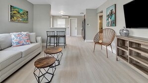 A modern living room with a sofa, two wooden tables, a wicker chair, and a TV on a wooden console. - Cozy 1BR Seaside, Steps to Hollywood Beach & More (Hollywood)