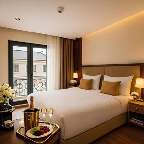 A hotel room with a large bed, a bedside table with a lamp, a tray with fruits and champagne, and a view of buildings through a window.