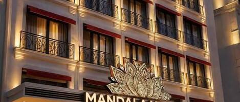 A multi-story hotel with balconies, a Christmas tree, and a sign reading 'MANDALA HOTEL KARAKOV'.