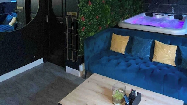 A modern living room with a blue sofa, a wooden coffee table, a green wall with plants, a black circular mirror, and a ceiling fan.