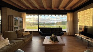 A modern living room with a wooden ceiling, large windows, a sofa, a coffee table with a floral arrangement, and a TV on a wooden stand.