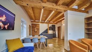 Un interior moderno con techo de madera, un comedor con mesa y sillas, una cocina al fondo, y un televisor mostrando un fuego.