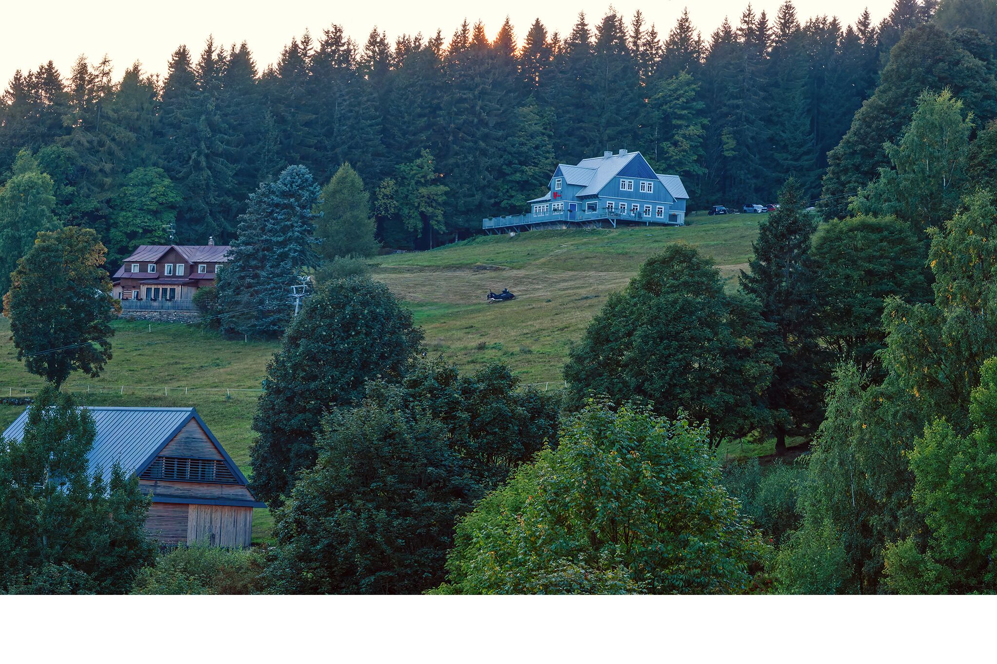 Une grande maison avec un toit en pente et une cour clôturée, entourée d’une forêt dense.