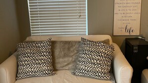 Un canapé en cuir blanc agrémenté de coussins à motifs, une fenêtre avec des stores et un panneau mural portant une citation sur l’amour.
