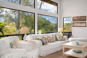 A modern living room with a white sofa, colorful pillows, a coffee table, and large windows offering a view of a lush garden and trees. - Serene Sandy Beach Golf Course Home (Honolulu)