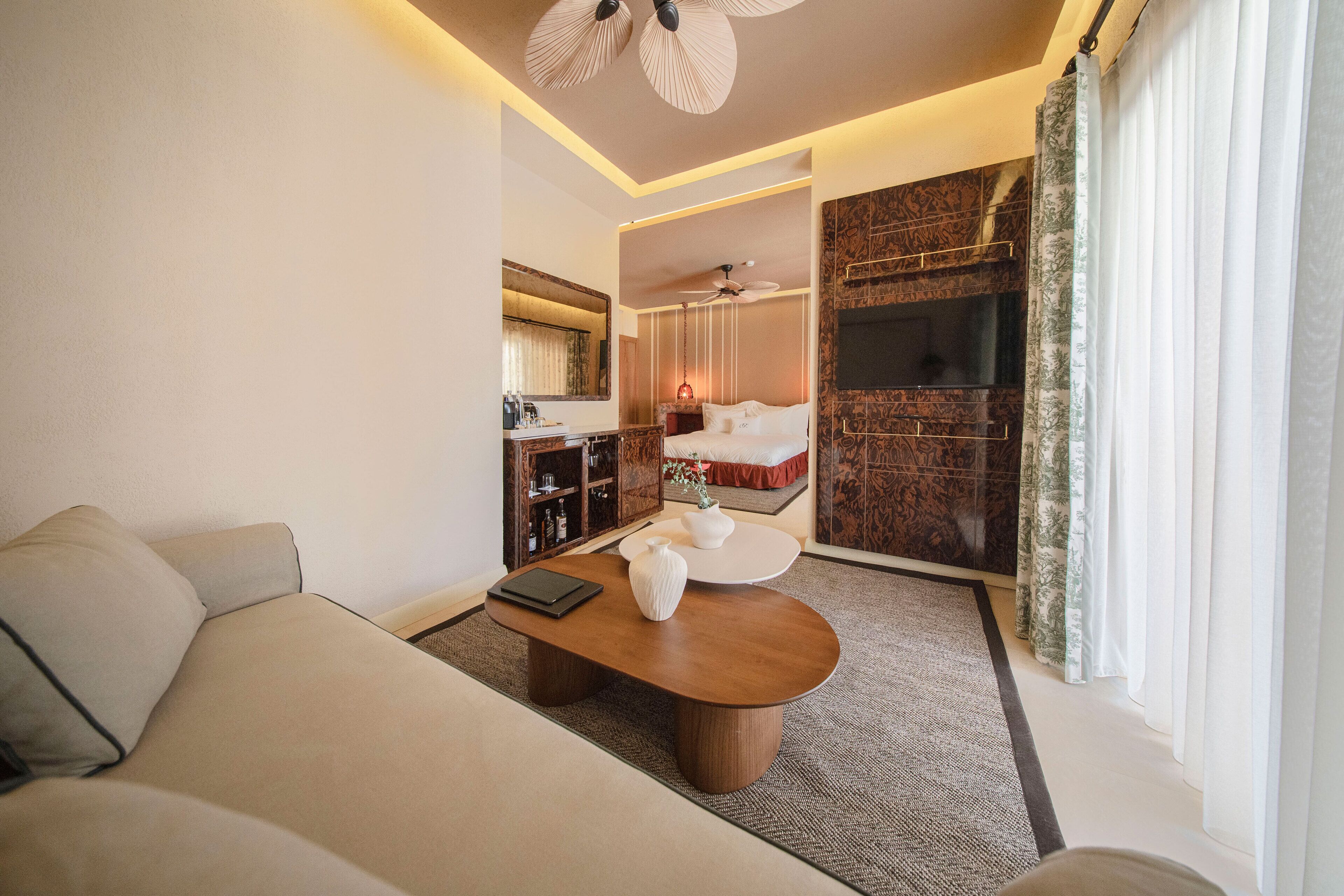 Ein modernes Hotelzimmer mit einer Couch, einem Holztisch, einer kleinen Küchenzeile, einem Bett und einem großen Fenster mit Vorhängen.