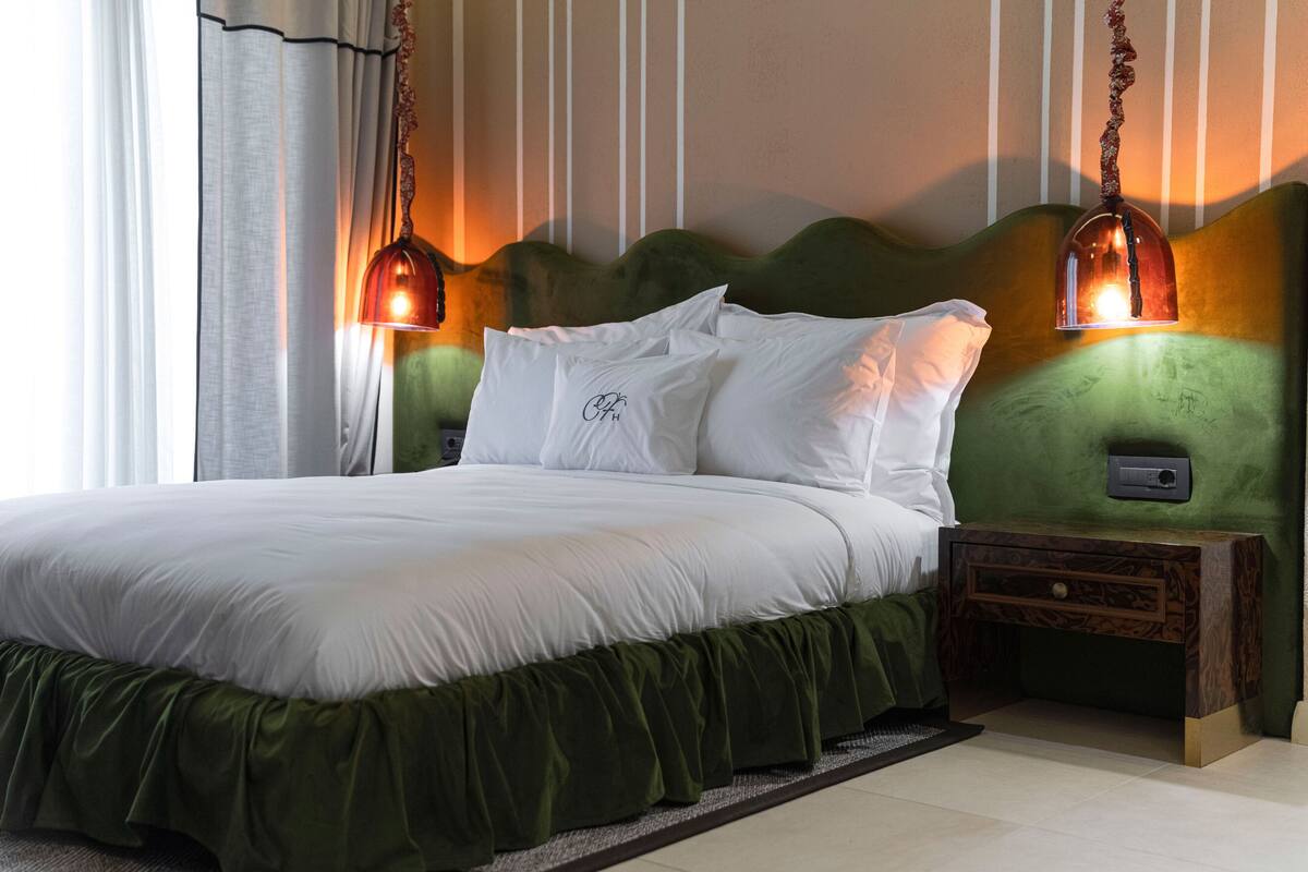 A neatly made bed with white linens, a green upholstered headboard, and two wall-mounted lamps.