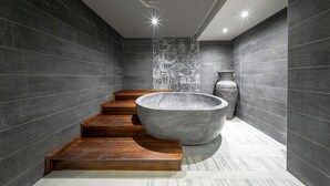A modern bathroom with dark tiled walls, a large stone basin, wooden steps, and a decorative wall panel. - Hotel Casa Fontana (Cesme)