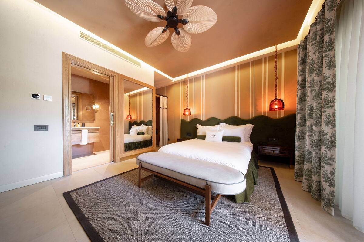 A modern bedroom with a large bed, a ceiling fan, a mirror, and a bathroom visible through an open door.
