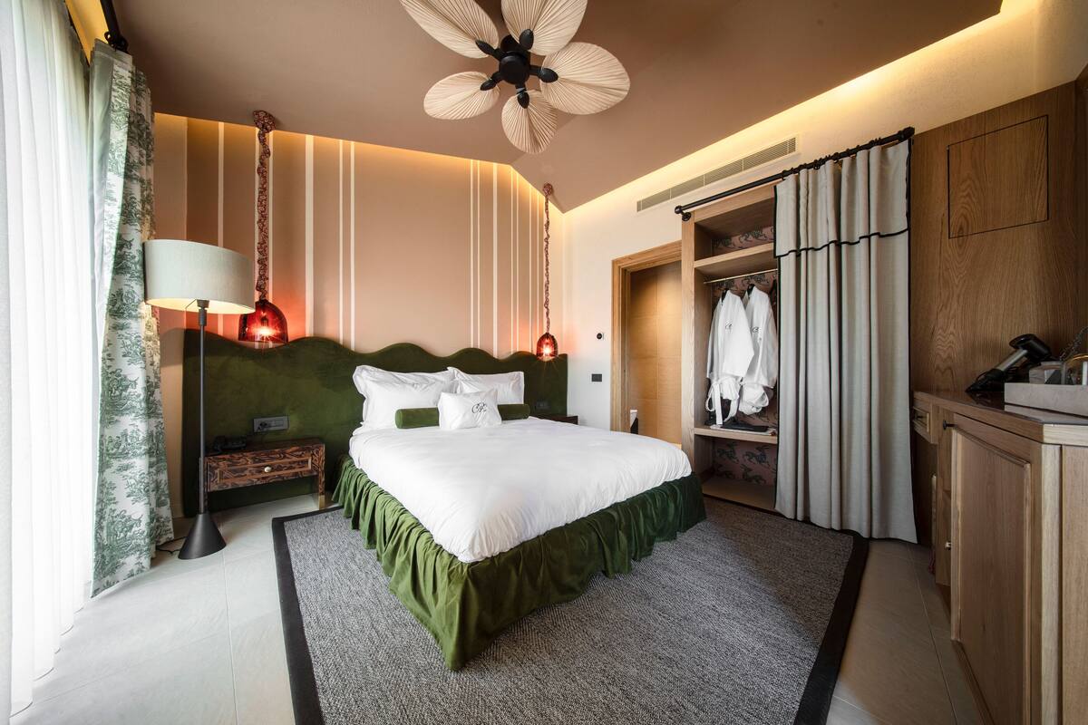 A hotel room with a large bed, a bedside lamp, a ceiling fan, a wardrobe, and a small table with a lamp.