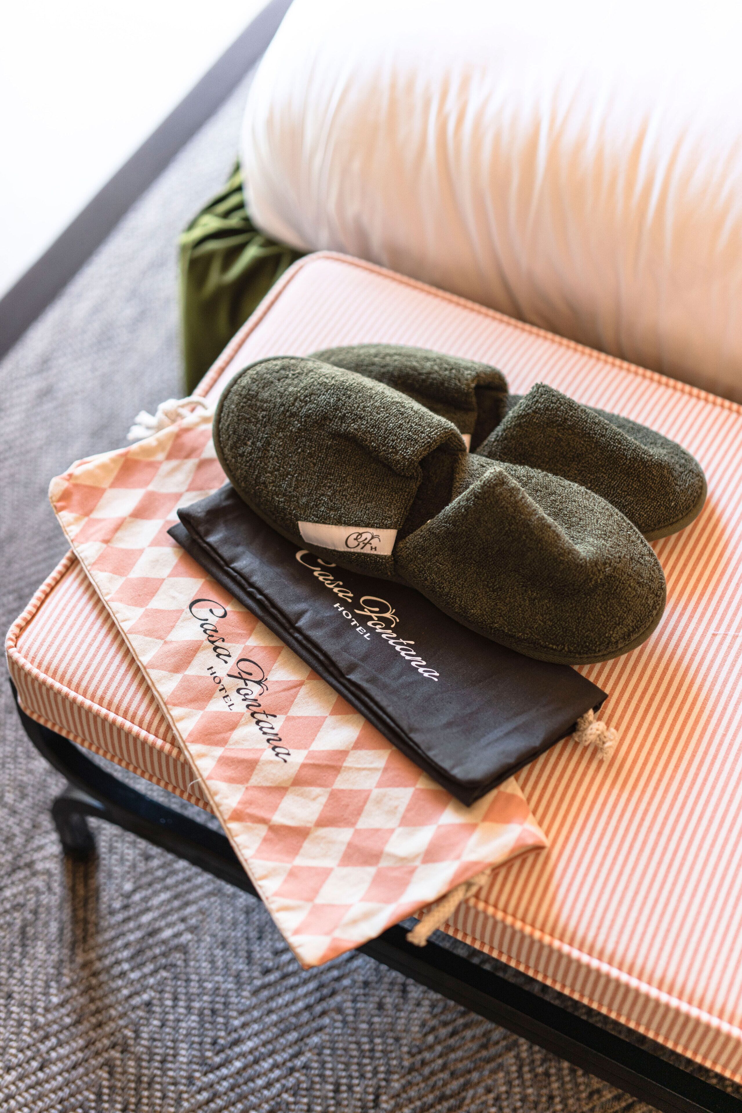 A neatly arranged hotel room with a checkered pillow, a pair of slippers, and a black bag on a bed.