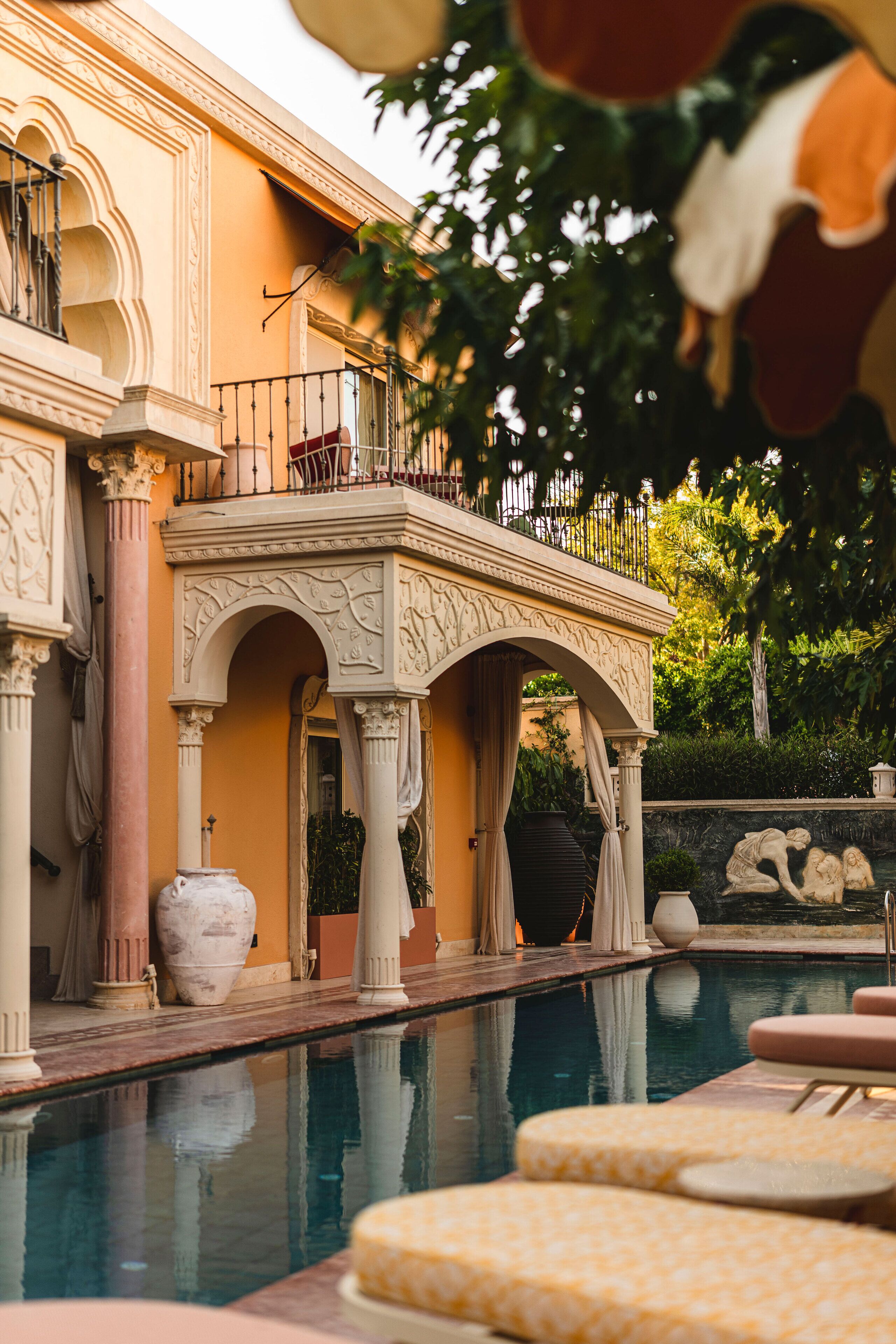Uma área de piscina com guarda-corpo, um terraço coberto com colunas e uma estátua ao fundo.