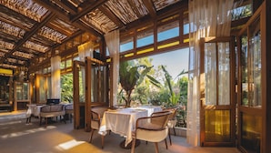 A dining area with a table set for four, surrounded by wicker chairs, large windows, and a view of greenery outside. - Hotel Casa Fontana (Cesme)