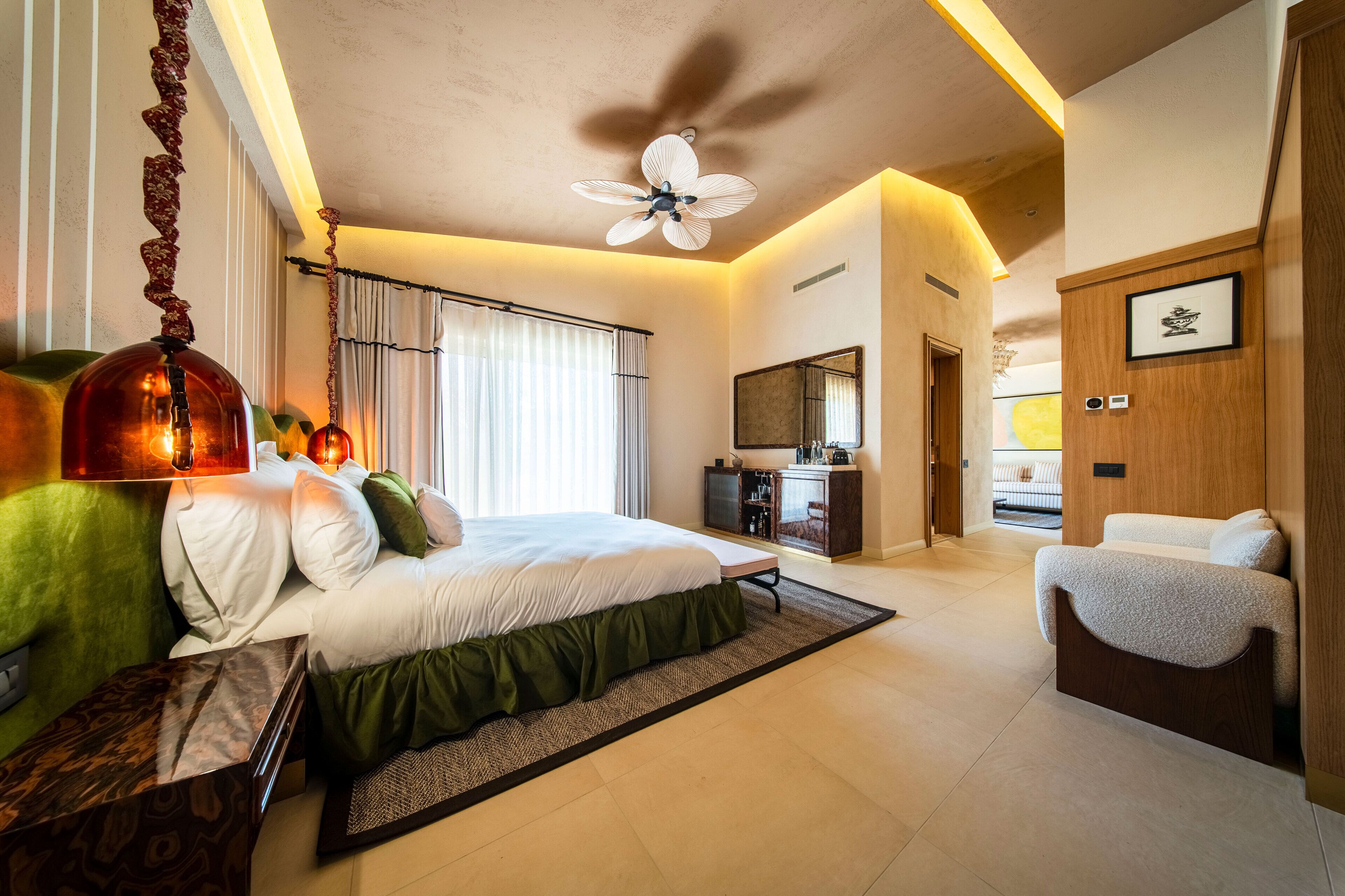 Quarto de hotel moderno com cama grande, ventilador de teto, escrivaninha e vista para o corredor.