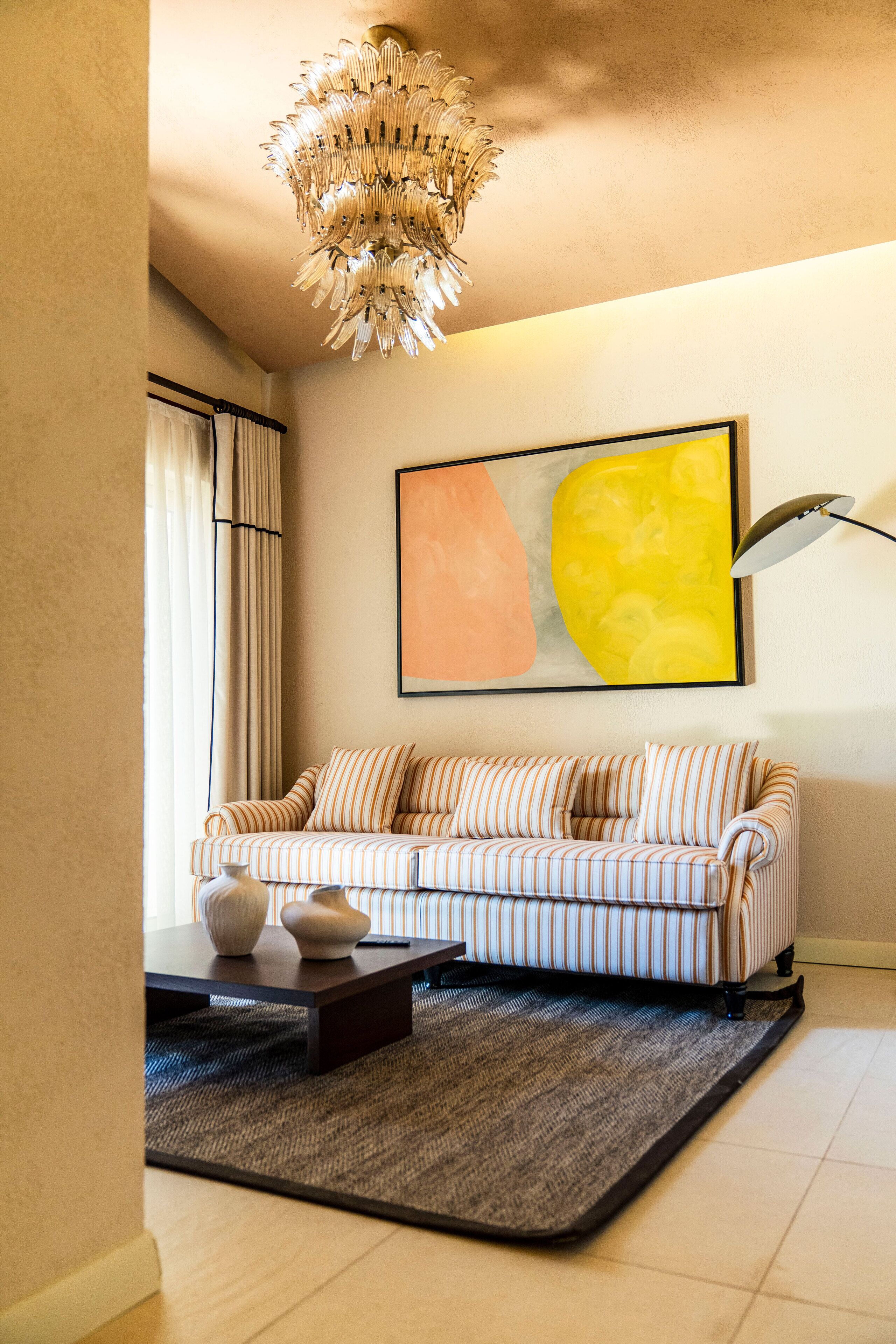 A living room with a striped sofa, a modern chandelier, a large abstract painting, and a floor lamp.