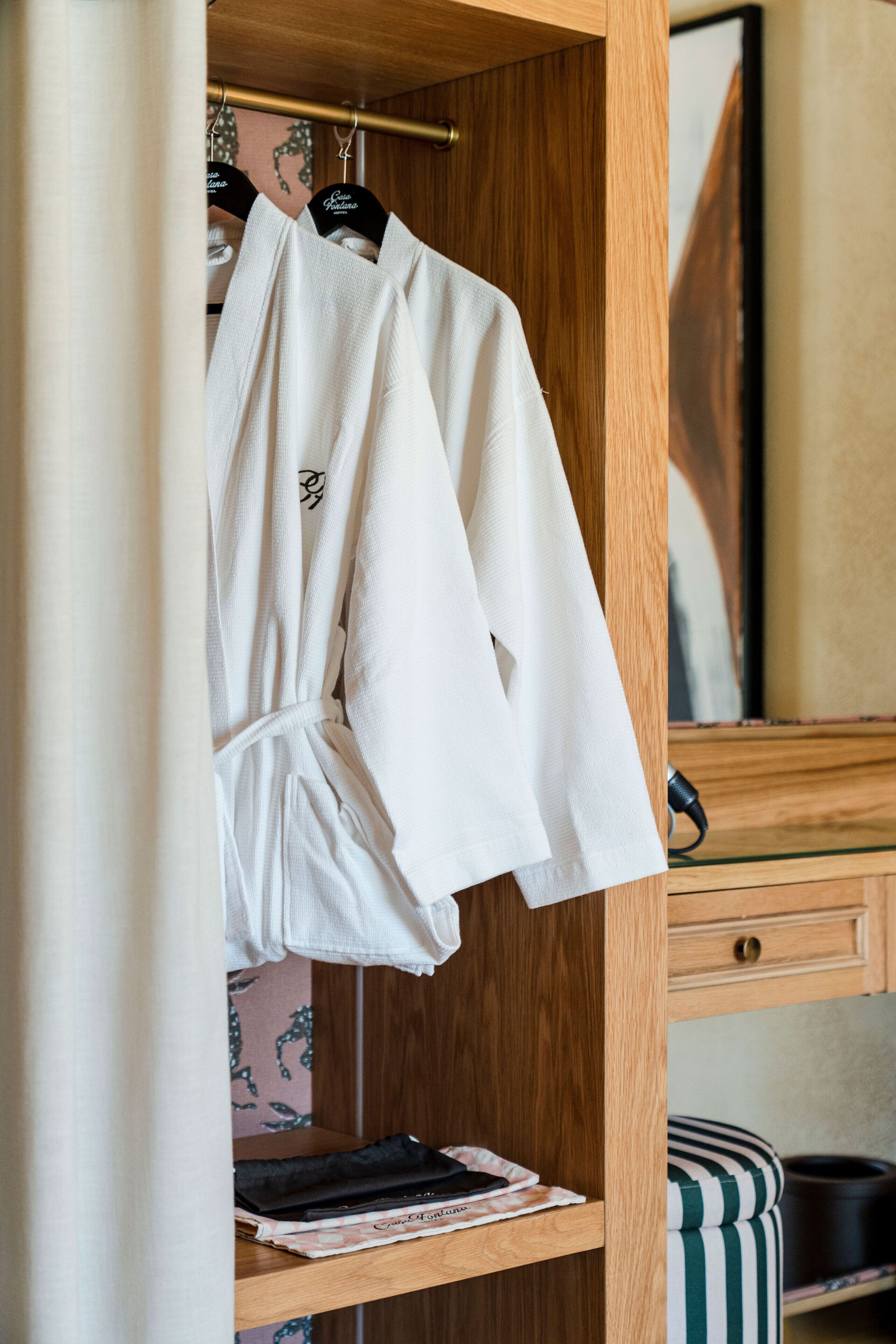 A wooden wardrobe with a robe hanging on a rod, a mirror, and a shelf with a book and a pair of slippers.