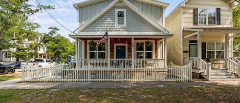 A two-story house with a front porch, a white picket fence, and an American flag.