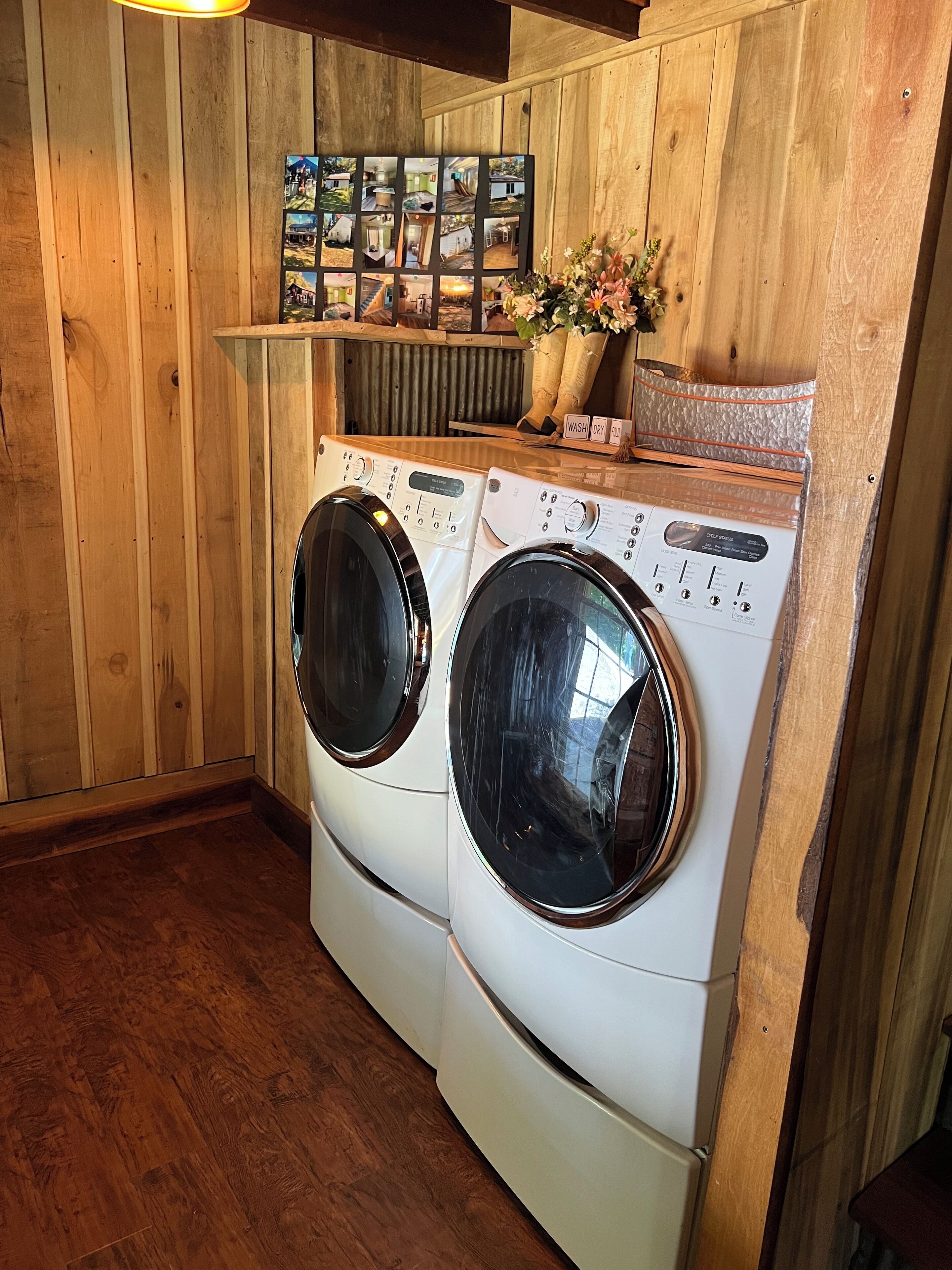 A laundry room with two front-loading washing machines, wooden walls, a shelf with framed photos, and a small plant.