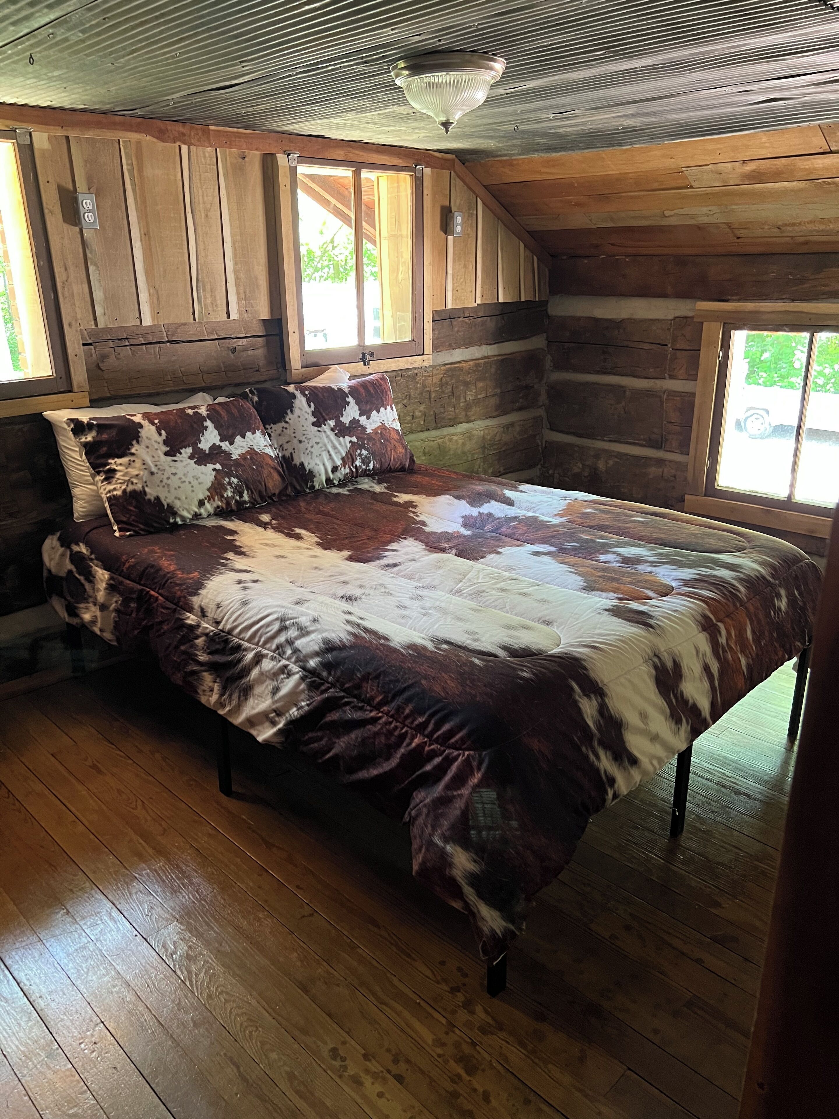 Un dormitorio tipo cabaña de madera con una cama de madera, un ventilador de techo y una ventana con vista a la vegetación.