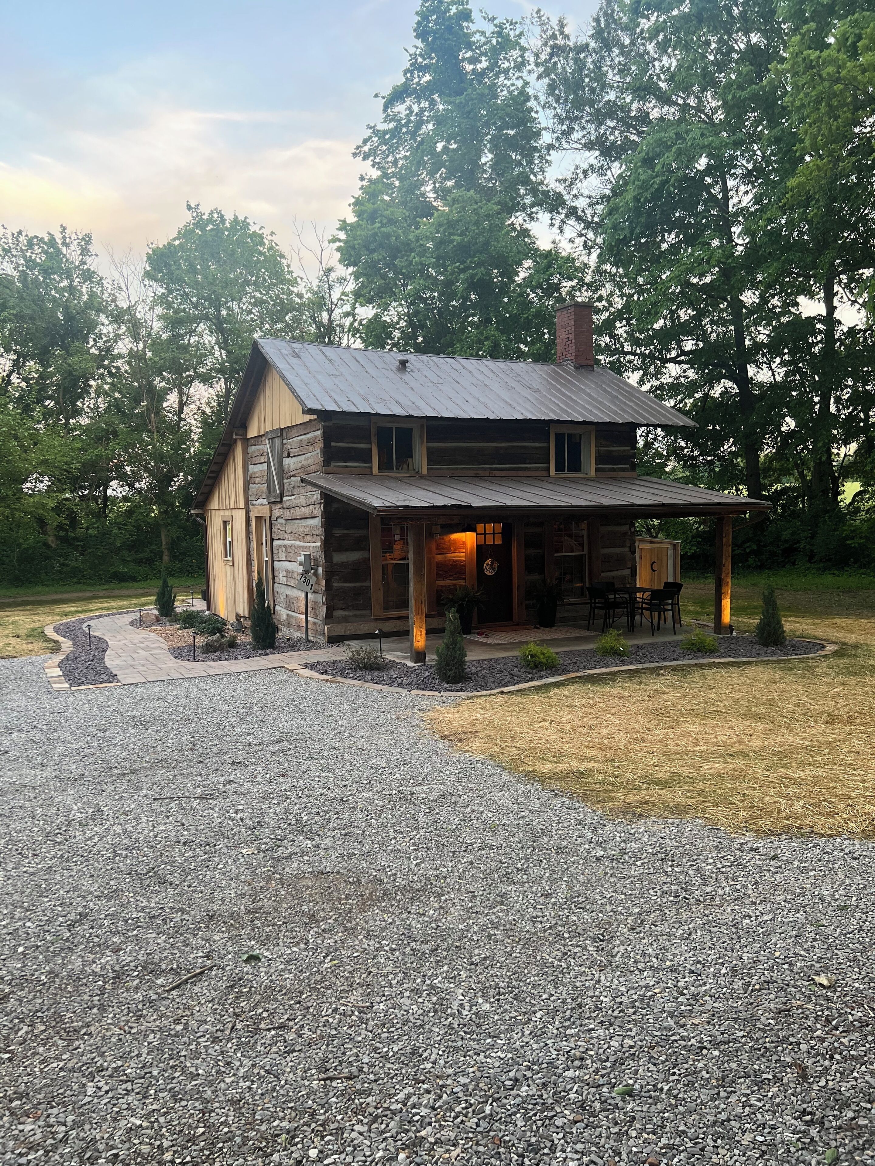 A rustic log cabin with a stone foundation, a porch with outdoor seating, and a gravel driveway.
