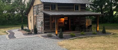 A rustic log cabin with a stone foundation, a porch with outdoor seating, and a gravel driveway.