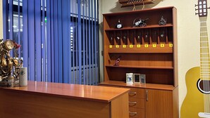 Ein Zimmer mit Schreibtisch aus Holz, einer Bücherwand mit Nummern und einer Gitarre, sowie einem Fenster mit blauen Jalousien.