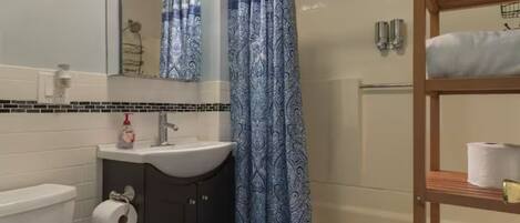 A bathroom with a white toilet, a sink with a mirror above it, a shower curtain, and a wooden shelf with a roll of toilet paper.