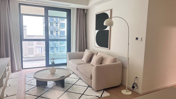 Un salon moderne avec un canapé, une table basse et un lampadaire. Une grande fenêtre, ornée de rideaux, offre une vue sur les bâtiments environnants.