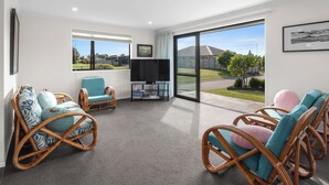 A spacious living room with wicker furniture, a flat-screen TV, and large windows offering a view of the outdoors.