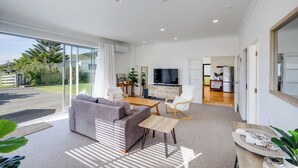 Ein geräumiges Wohnzimmer mit grauem Sofa, einem Holztisch und einem Fernseher. Eine Glastür ermöglicht den Zugang zu einem Balkon mit Blick auf Grünflächen und einem Zaun.