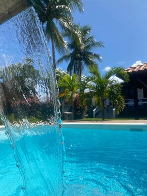 A clear view of a swimming pool with water cascading over a structure, surrounded by palm trees and a building with a tiled roof. - Solar dos Oliverias Serrambi by La Playa (Ipojuca)