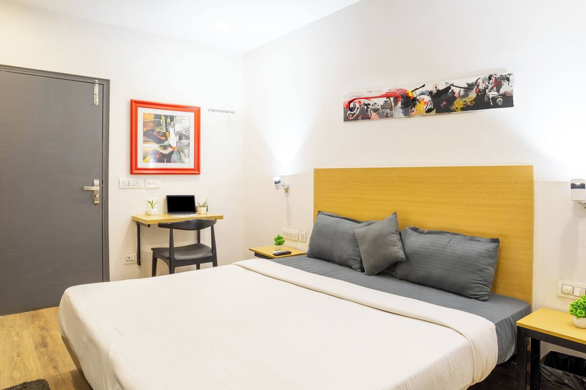 A hotel room with a large bed, a desk, a chair, a small table with a plant, and artwork on the walls.