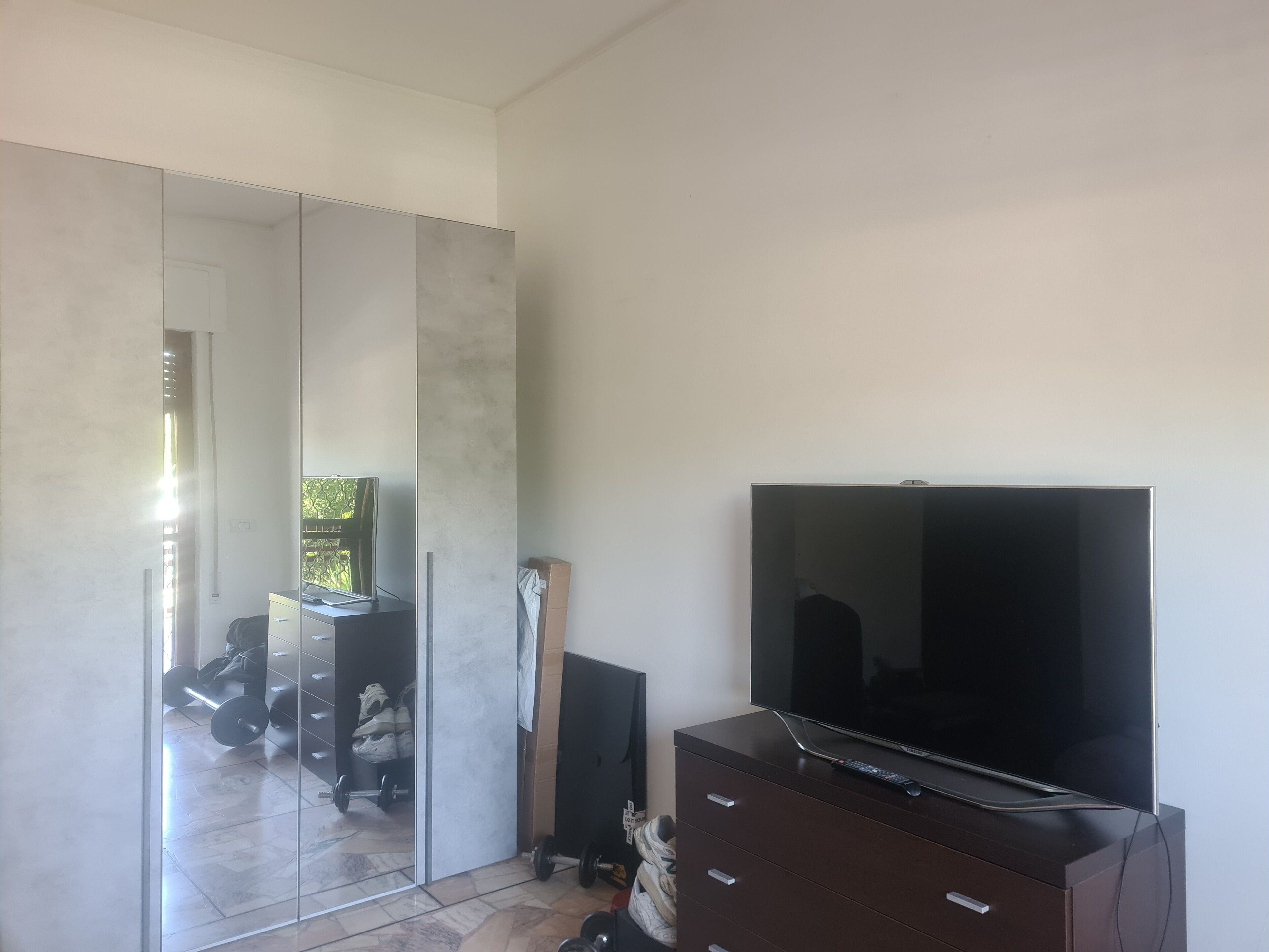 Una camera moderna con un grande specchio, una TV a schermo piatto su un mobile in legno e una porta scorrevole in vetro che dà su un balcone con attrezzature per l'esercizio fisico.
