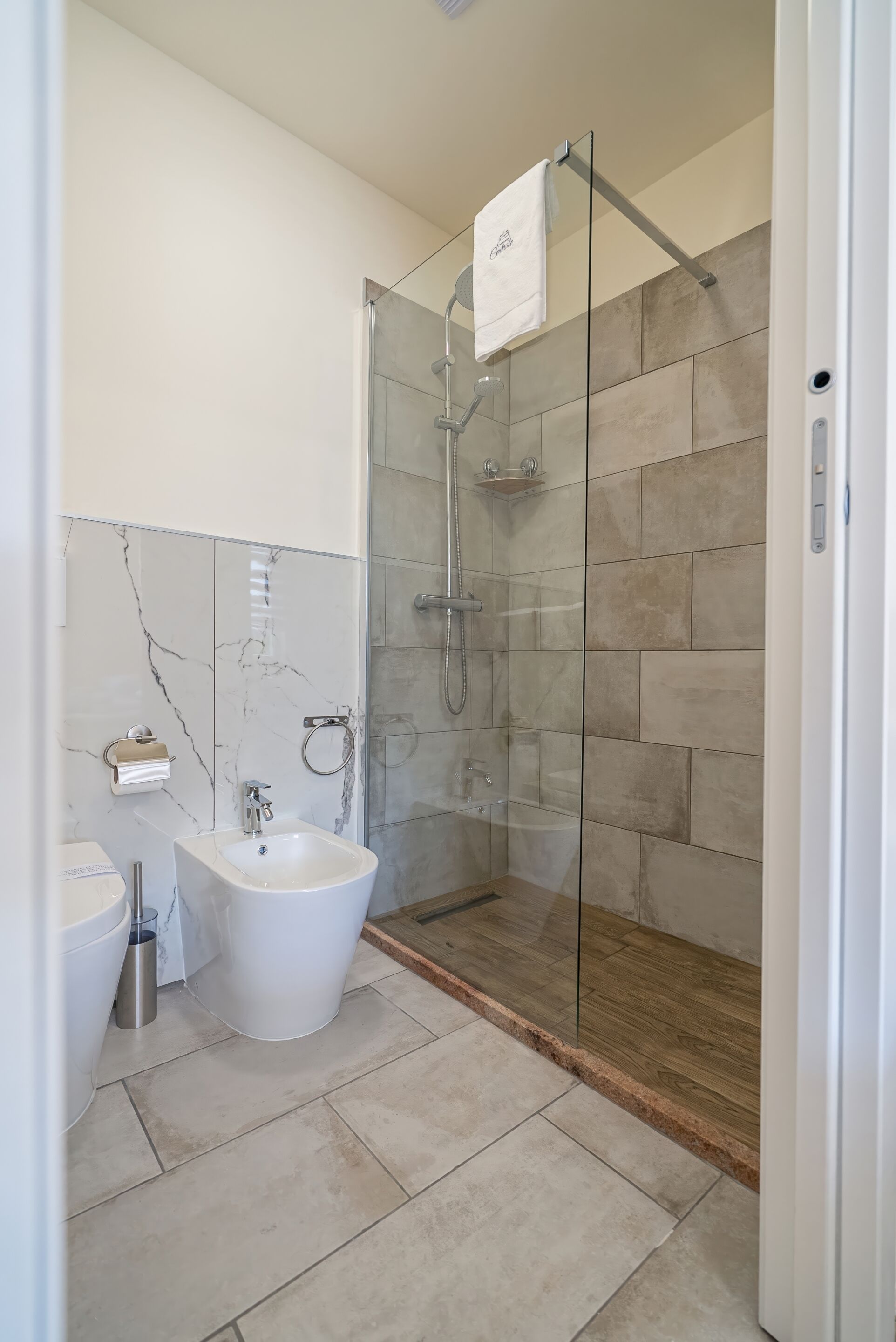 Een moderne badkamer met een douchecabine van glas, een witte wastafel en een wand betegeld met marmer.