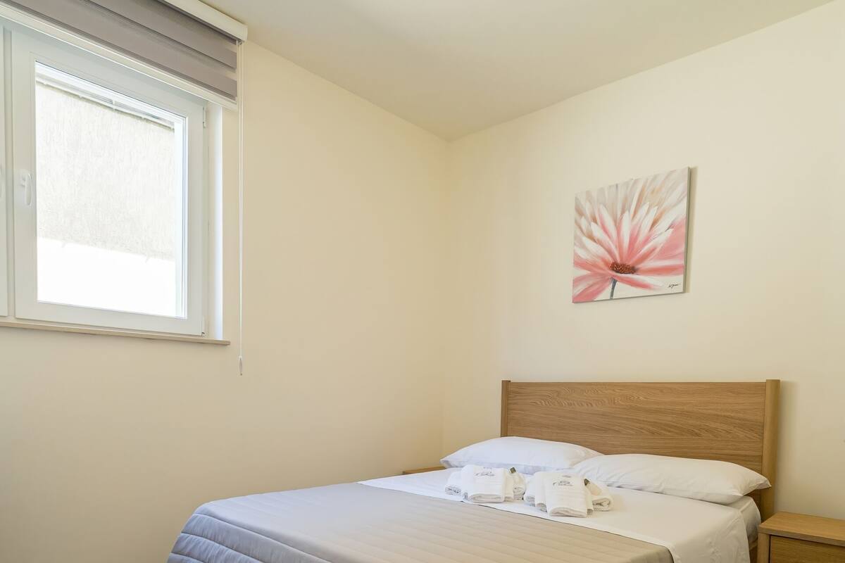 A bedroom with a bed, a nightstand, a window with blinds, and a wall-mounted artwork.