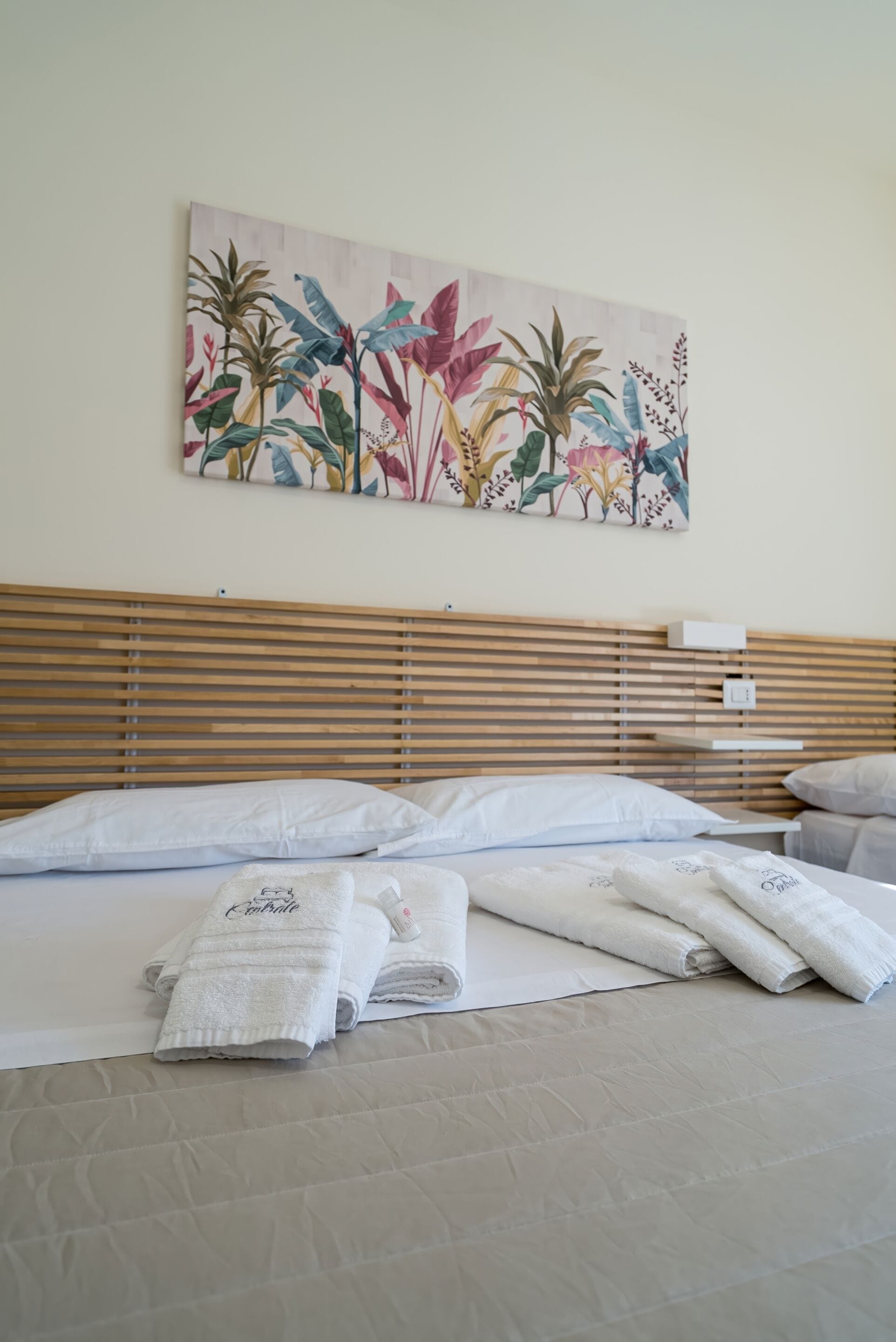 A neatly made bed with white linens and a folded towel, a wooden headboard, and a colorful floral painting on the wall.