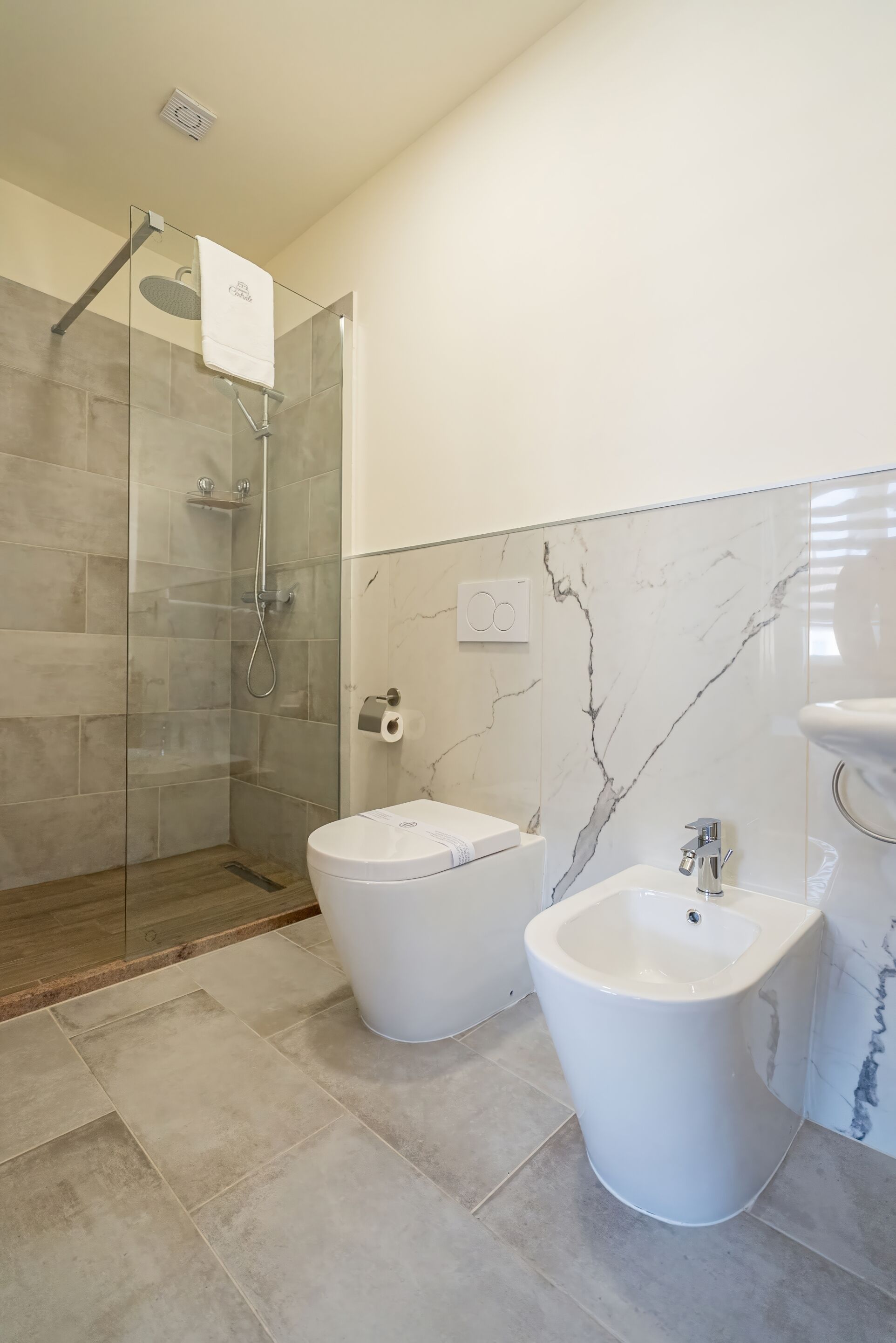 Un baño moderno con ducha envuelta en cristal, dos bidés blancos, un lavamanos blanco y paredes con diseño de mármol.