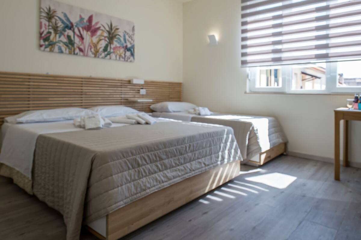 A bedroom with two beds, a wooden headboard, a window with blinds, and a painting on the wall.