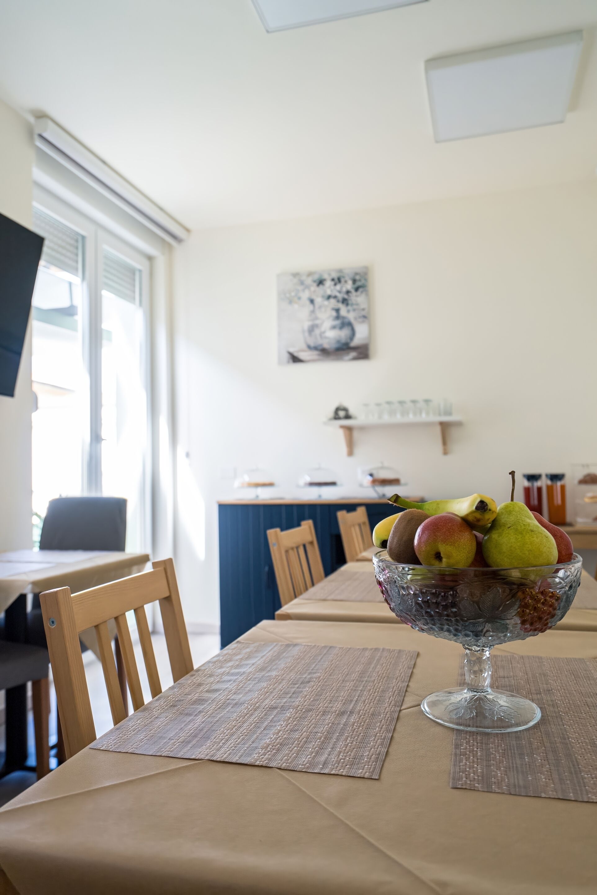 A dining table with a glass bowl of fruit, a wooden chair, and a view of a kitchen area with a window and a painting on the wall.