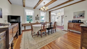 A dining area with a wooden table, chairs, and a chandelier. There is a television on a stand and a refrigerator in the background.