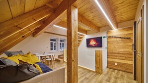 Ein Schlafzimmer im Loft-Stil mit schrägverdeckter Decke, Holzbalken, einem Bett mit Kissen, einem an der Wand montierten Fernseher und einer Treppe, die in einen anderen Raum führt.