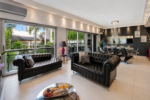 Un salon moderne avec deux canapés en cuir noir, un coin repas comprenant une table et des chaises, et de grandes fenêtres offrant une vue sur la verdure.