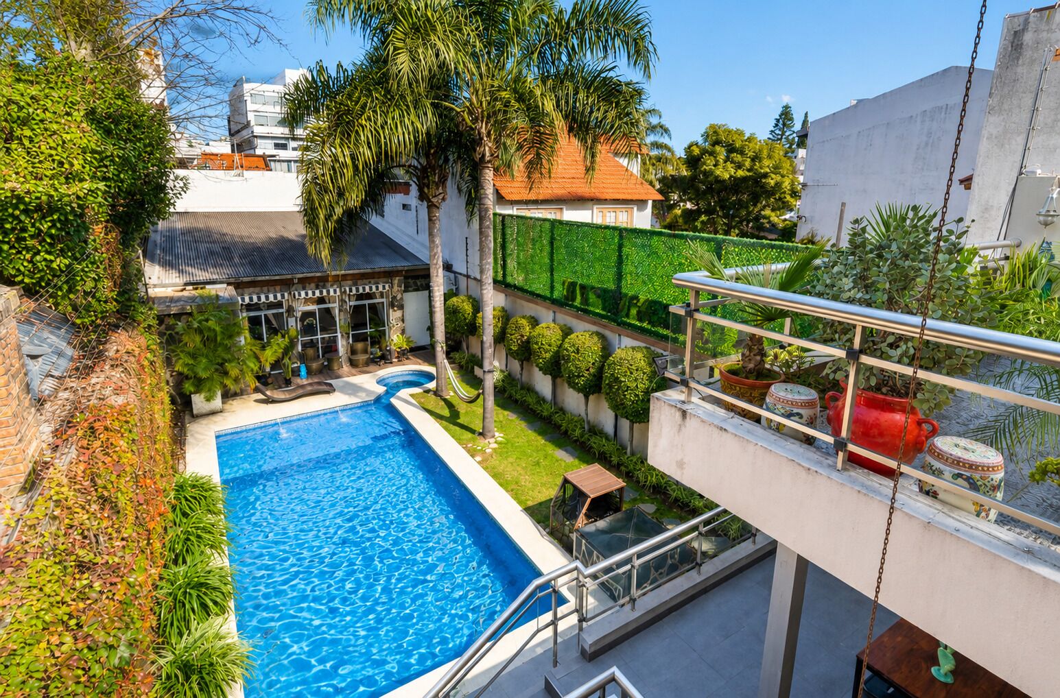 A rooftop swimming pool with a glass balustrade, surrounded by lush greenery and palm trees.