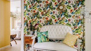 Una habitación con papel tapiz floral, un sofá blanco con tapicería y cojines estampados, una mesita de madera y una araña.