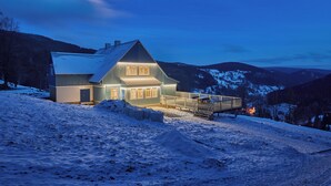 Een modern huis met een ruime veranda en een overdekt terras, omgeven door besneeuwde heuvels en een bergketen op de achtergrond.